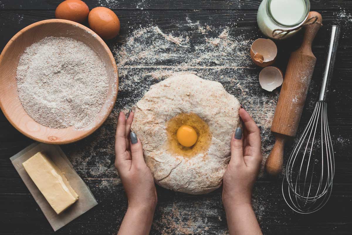 Hands holding dough with an egg well, surrounded by flour, a bowl of flour, eggs, butter, a rolling pin, and a whisk.